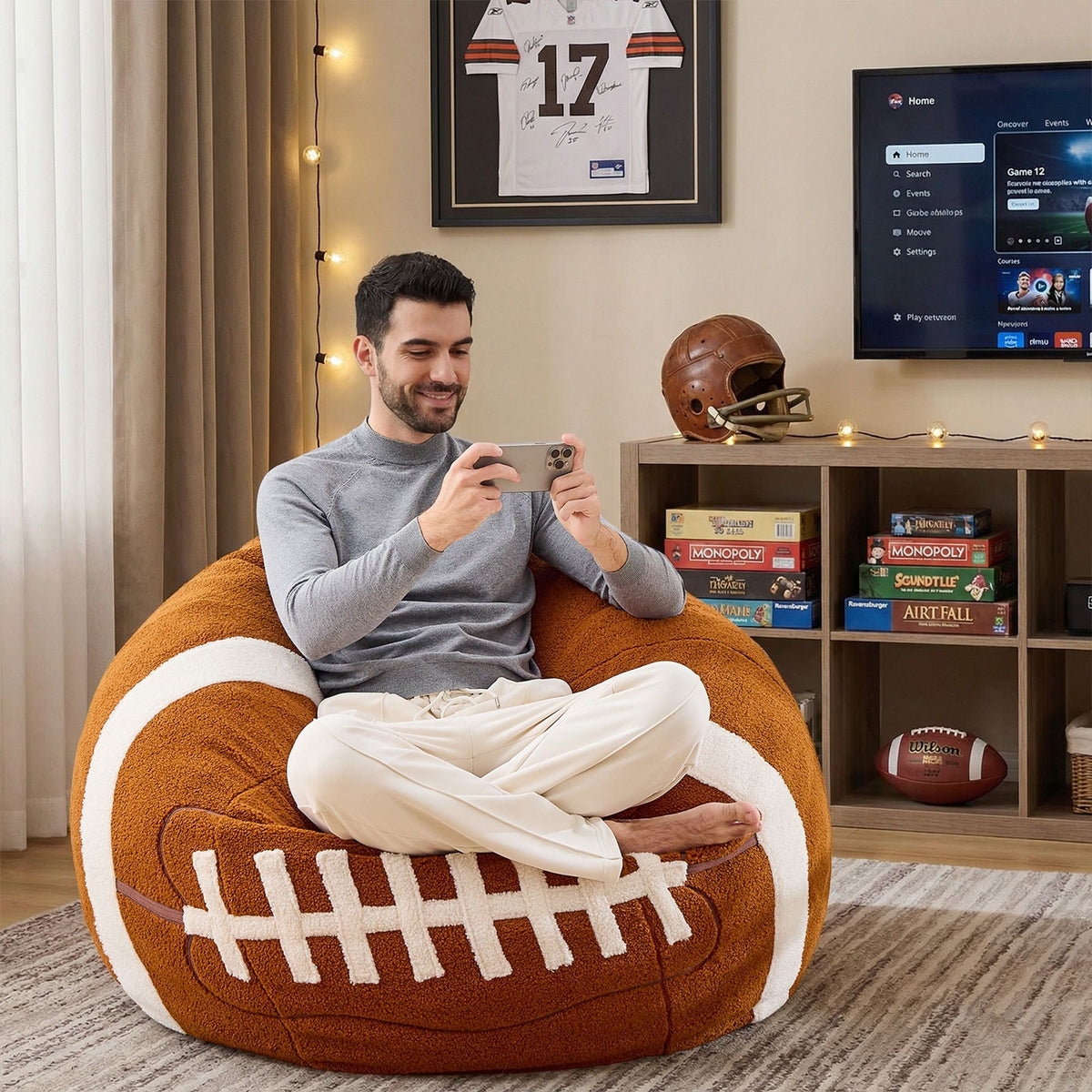 A man is sitting comfortably on the Giant Rugby Plush Sofa, playing with his mobile phone.