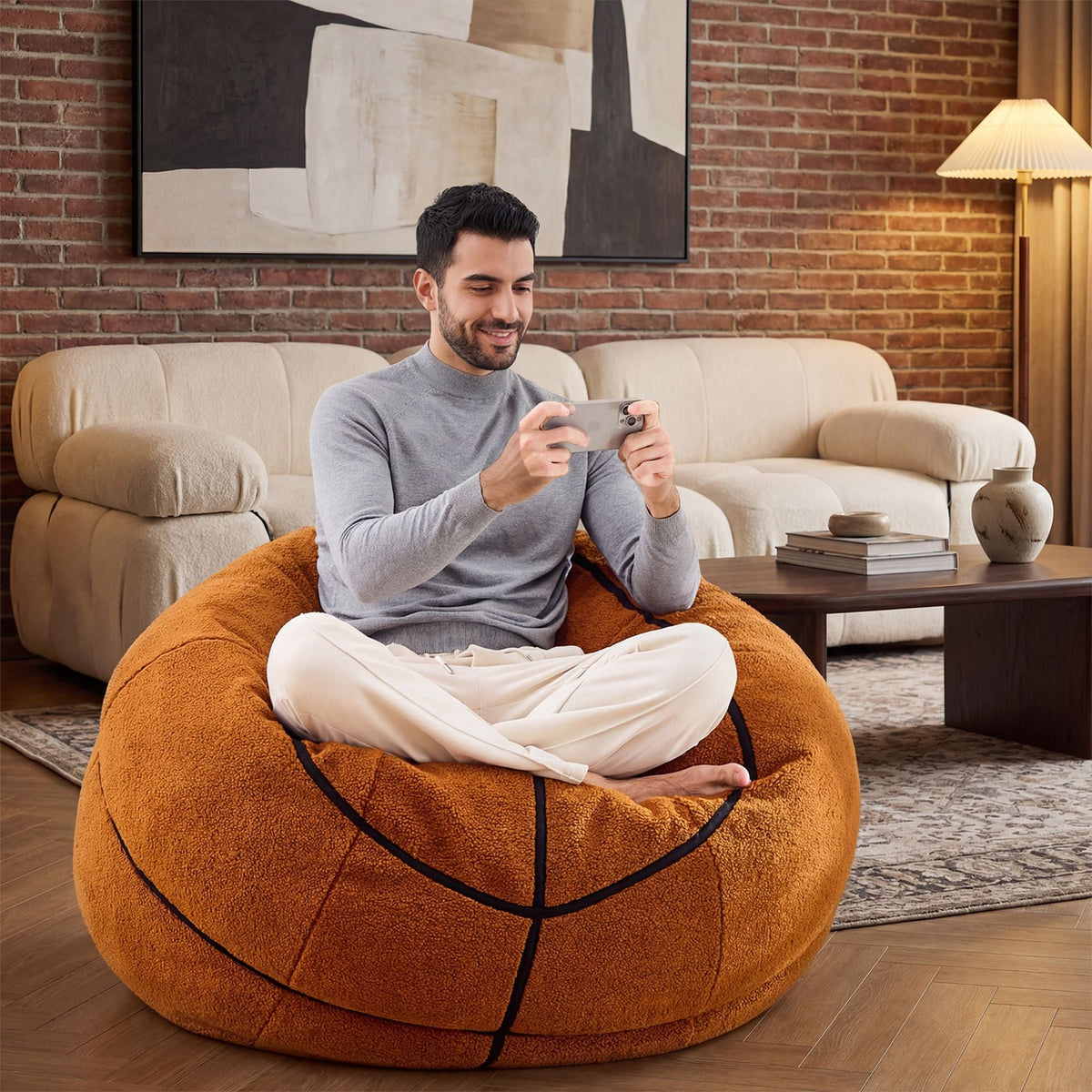 A man is sitting comfortably on the Giant Basketball Plush Sofa, playing with his mobile phone.