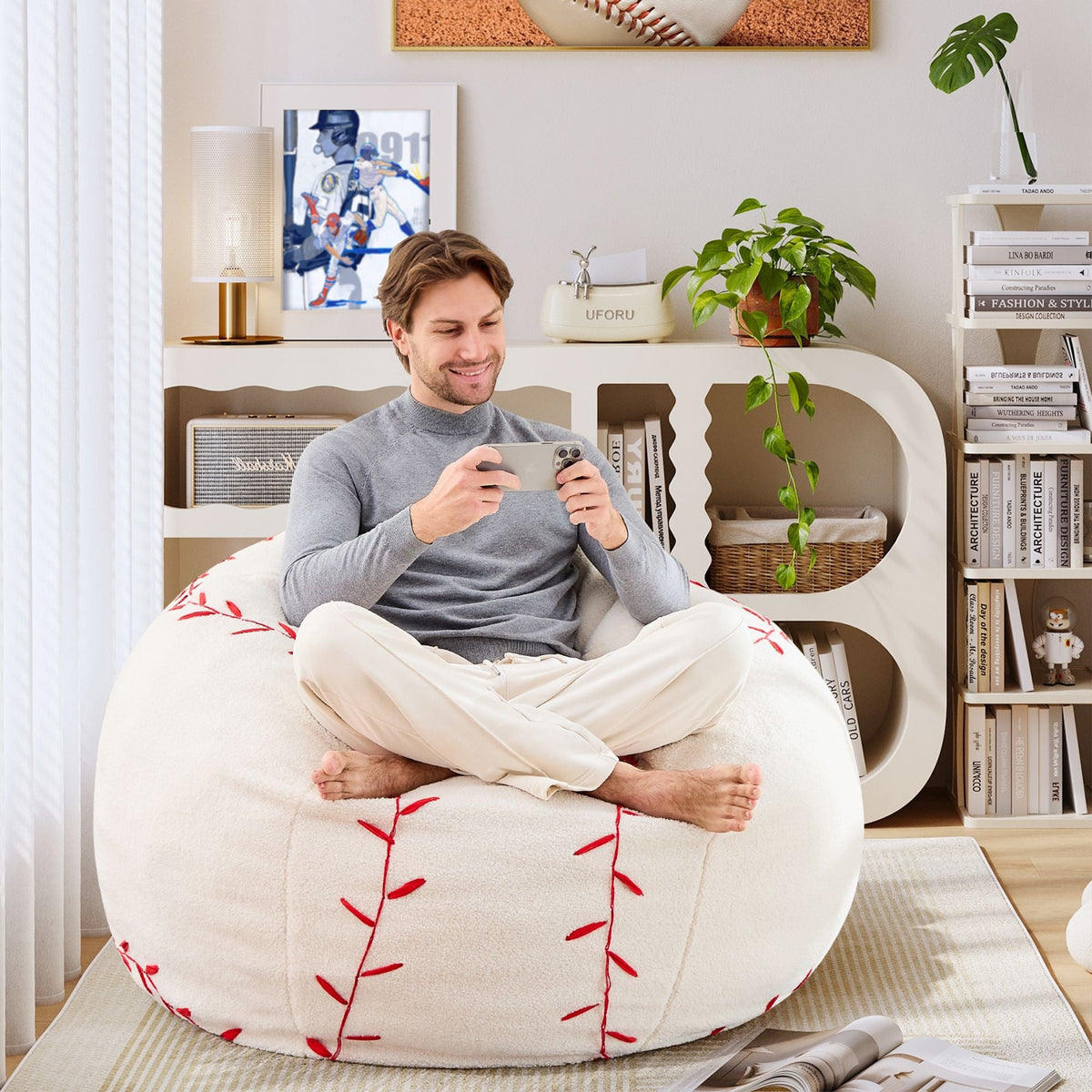 A man is sitting comfortably on the Giant Baseball Plush Sofa, playing with his mobile phone.